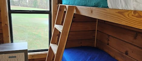 A bunk bed room with a wooden ladder, a small desk, and a window with a view of a grassy area.