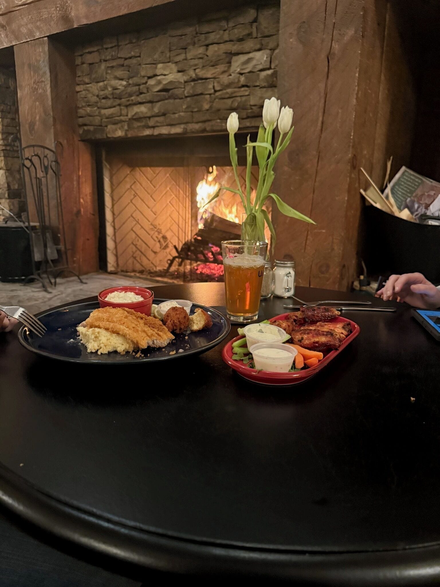 Una mesa con comida que incluye pollo empanizado, ensalada y una jarra de cerveza, frente a una chimenea de piedra con fuego encendido.