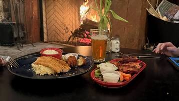A table with a meal consisting of breaded chicken, salad, and a glass of beer, set against a stone fireplace with a lit fire.