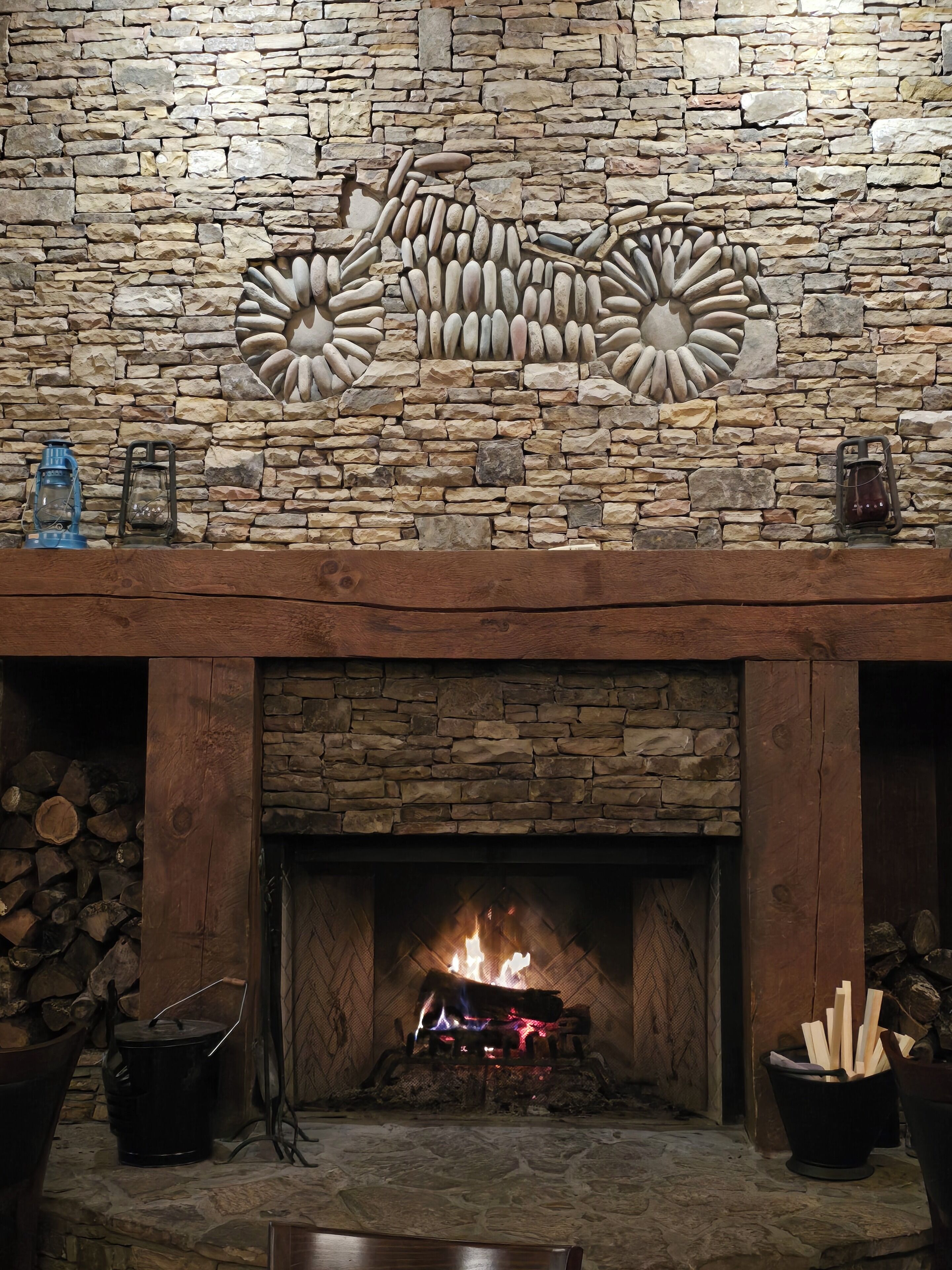 A stone fireplace with a fire burning, surrounded by logs and a rustic wooden mantel.
