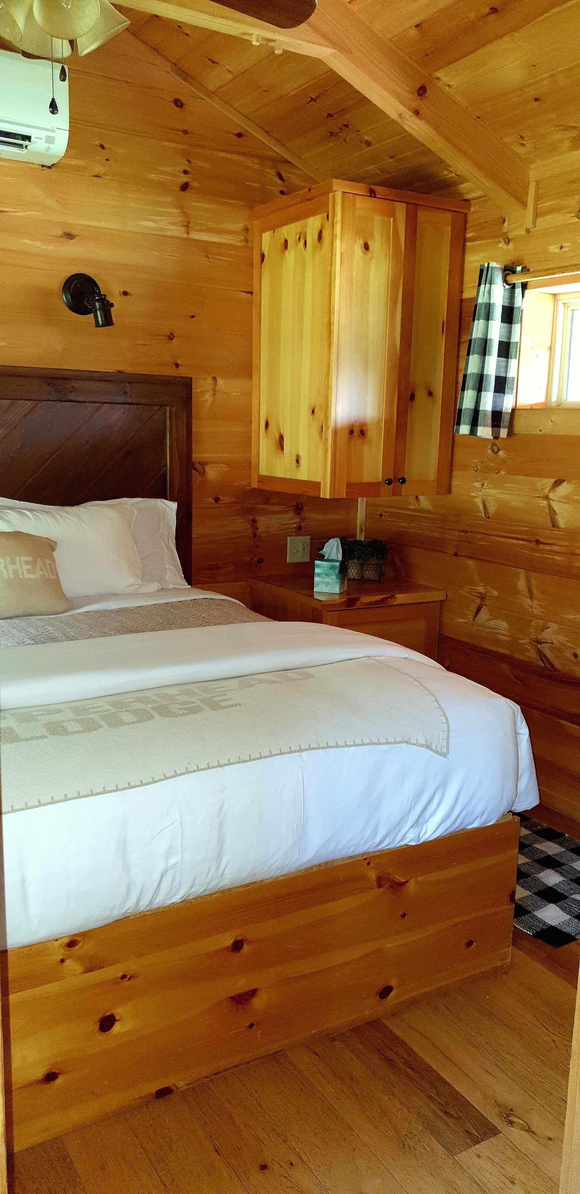 A wooden cabin bedroom with a bed, a headboard, a nightstand, and a window with curtains.