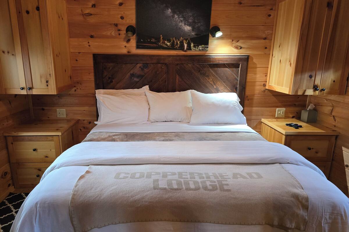 A wooden cabin-style bedroom with a bed, two bedside tables, a nightstand, a lamp, and a framed picture on the wall.