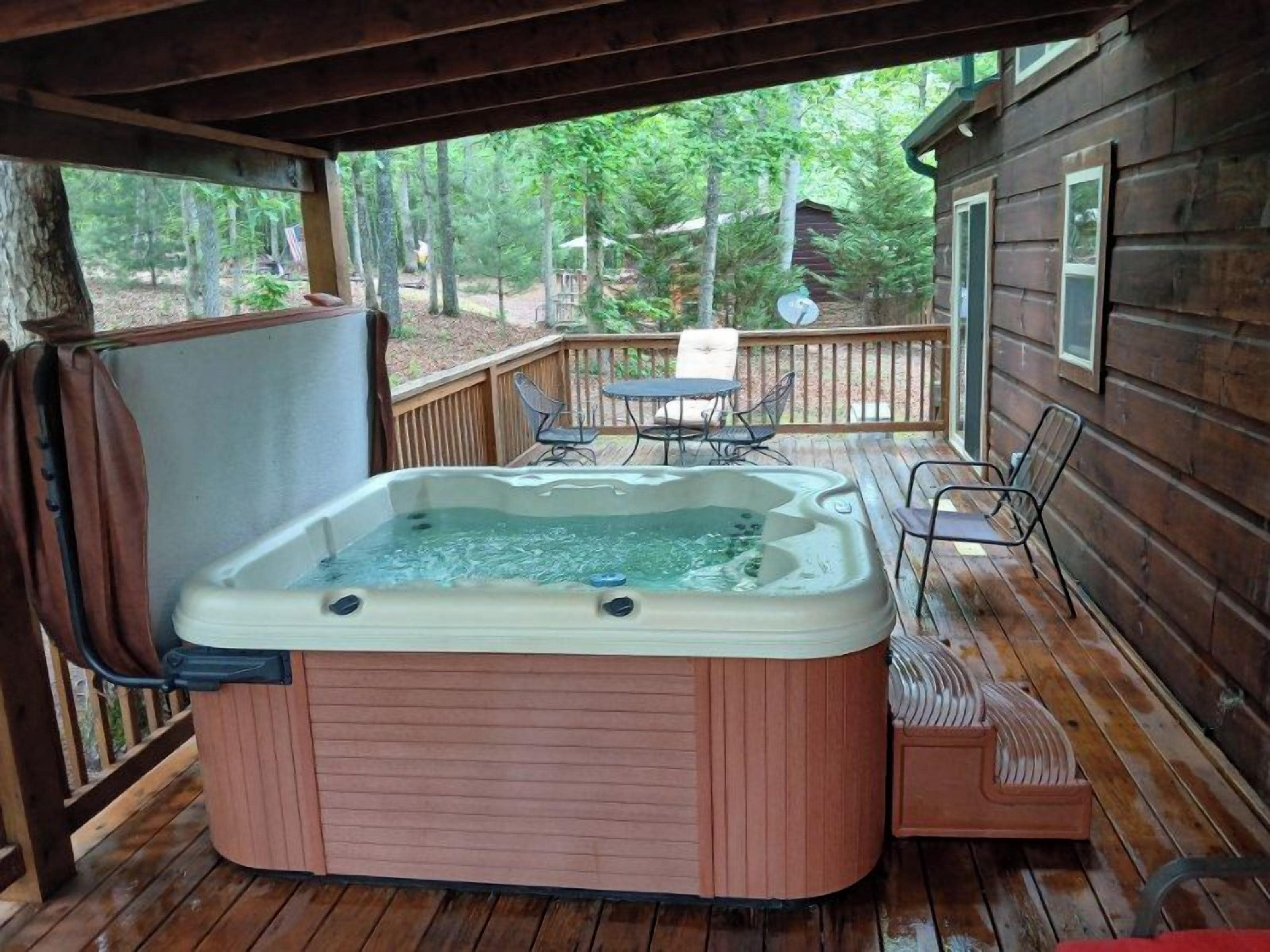 A hot tub on a wooden deck with a table and chairs, surrounded by trees.