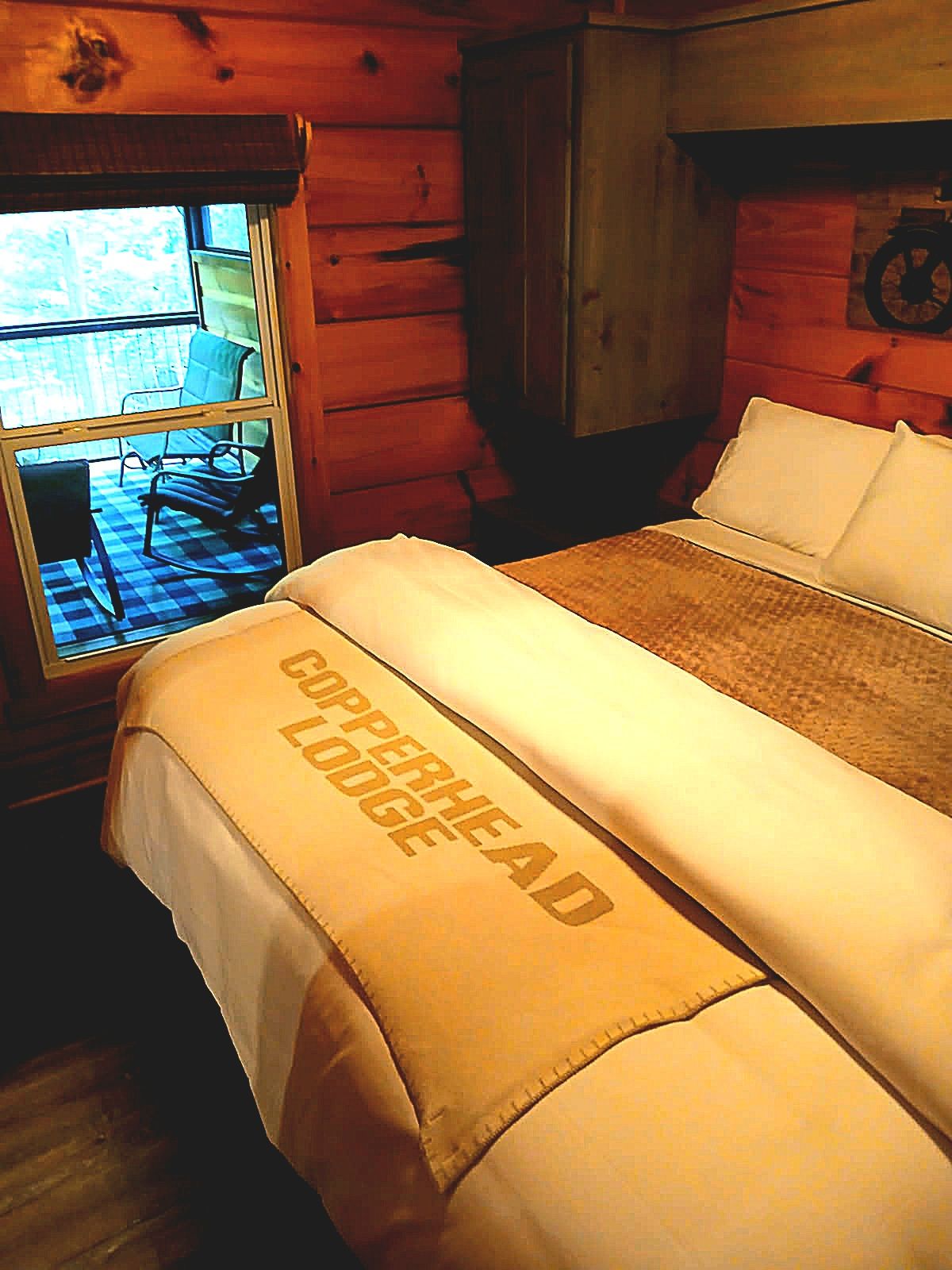 A bed with a blanket labeled 'Copperhead Lodge' in a log cabin room with a window overlooking a checkered floor and chairs outside.