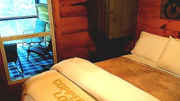 A bed with a blanket labeled 'Copperhead Lodge' in a log cabin room with a window overlooking a checkered floor and chairs outside.