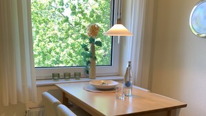 Zona pranzo con tavolo in legno, due sedie bianche, un vaso con fiori, una bottiglia e una finestra con tende.
