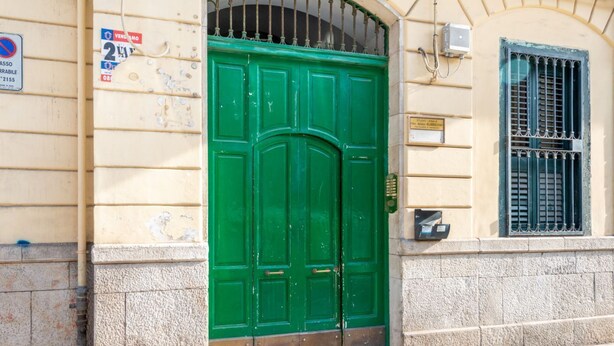 Una puerta doble verde con una reja metálica, un letrero encima y una ventana con barrotes a la derecha.