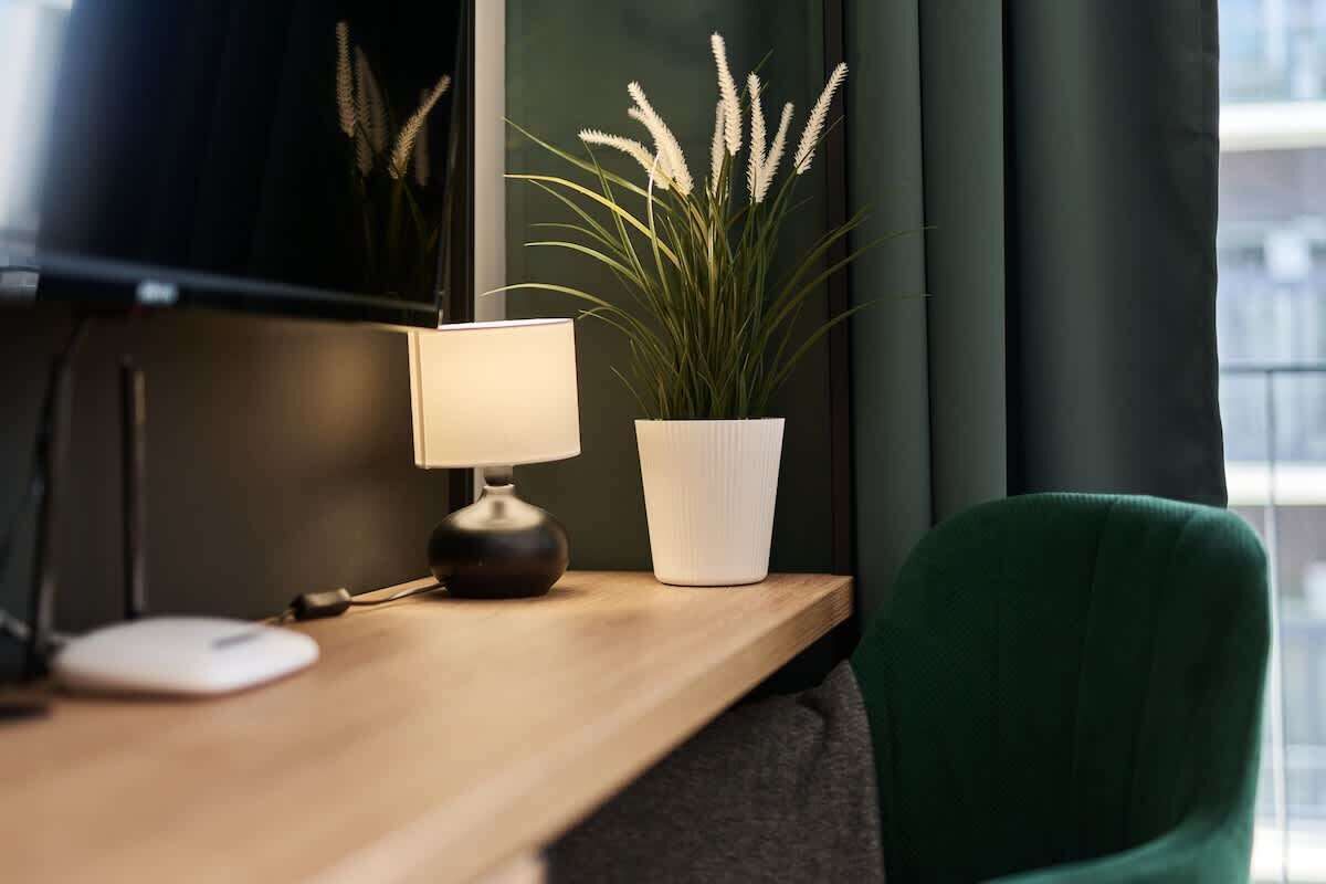 Una scrivania con una lampada, una pianta in vaso e una sedia verde in una stanza con una finestra e una parete scura.