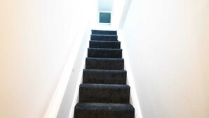 Un escalier étroit, recouvert de moquette, avec des murs blancs et une petite fenêtre en haut.