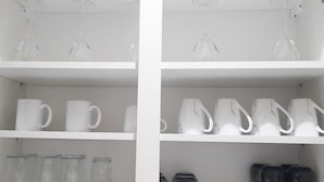 Une étagère blanche sur laquelle sont disposés divers objets en verre, notamment des tasses transparentes et blanches, ainsi que des bouteilles en verre.