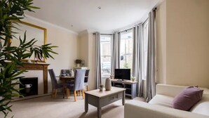 A living room with a sofa, a dining table with chairs, a TV, and a fireplace. - Spacious 2 Bed Flat Near High Street Kensington (London)