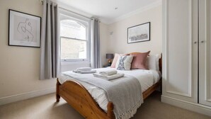 A bedroom with a wooden bed, a window with curtains, and two framed pictures on the walls. - Spacious 2 Bed Flat Near High Street Kensington (London)