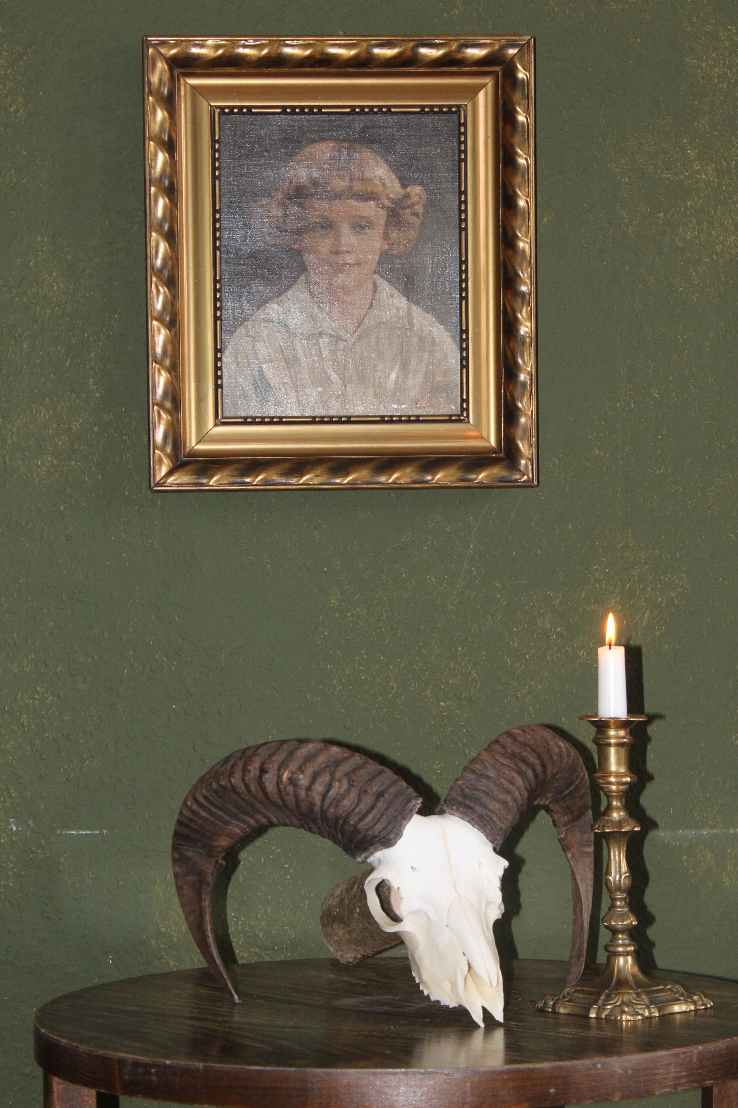 A framed portrait on a wall, a skull on a table, and a lit candle.