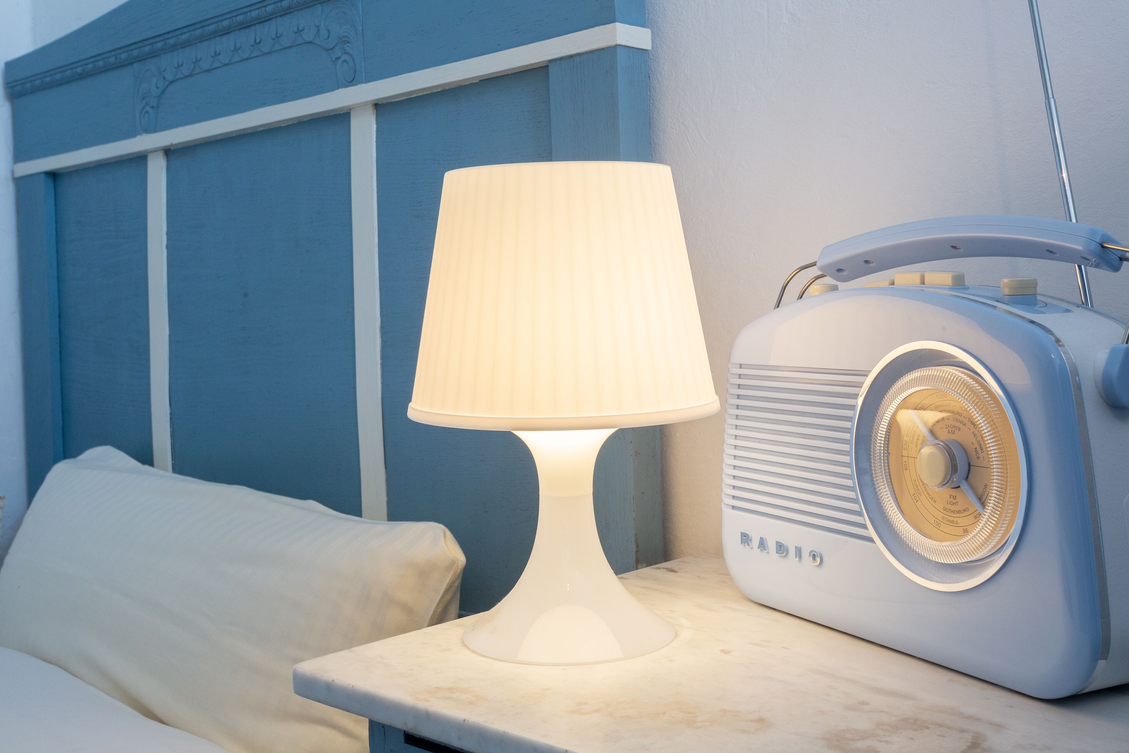 Una cama con un almohadón blanco y una lámpara de mesa blanca junto a una radio antigua.