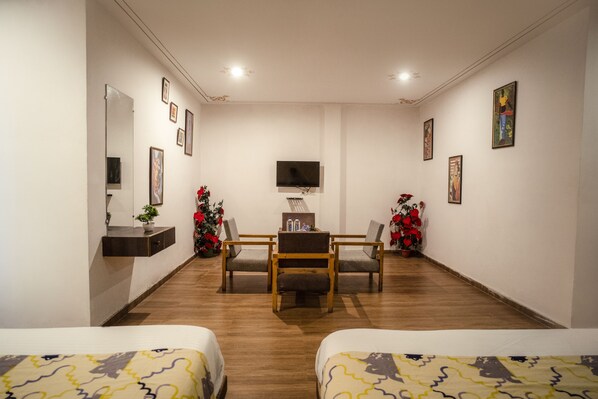 A hotel room with two beds, a desk with chairs, a TV, and wall-mounted artwork. - Tripli Hotels Arunoday Palace (Udaipur)