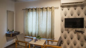 Une pièce dotée d’un téléviseur à écran plat fixé sur un mur orné de motifs, d’une table en bois avec des chaises, d’une petite plante en pot et d’une fenêtre avec des rideaux.