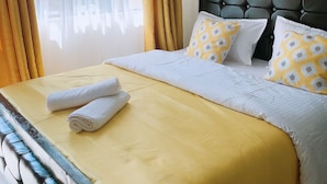 A neatly made bed with a yellow bedspread and pillows, a headboard, and a window with curtains. - Stay.Plus Hill View Studio Apartment (Nairobi)
