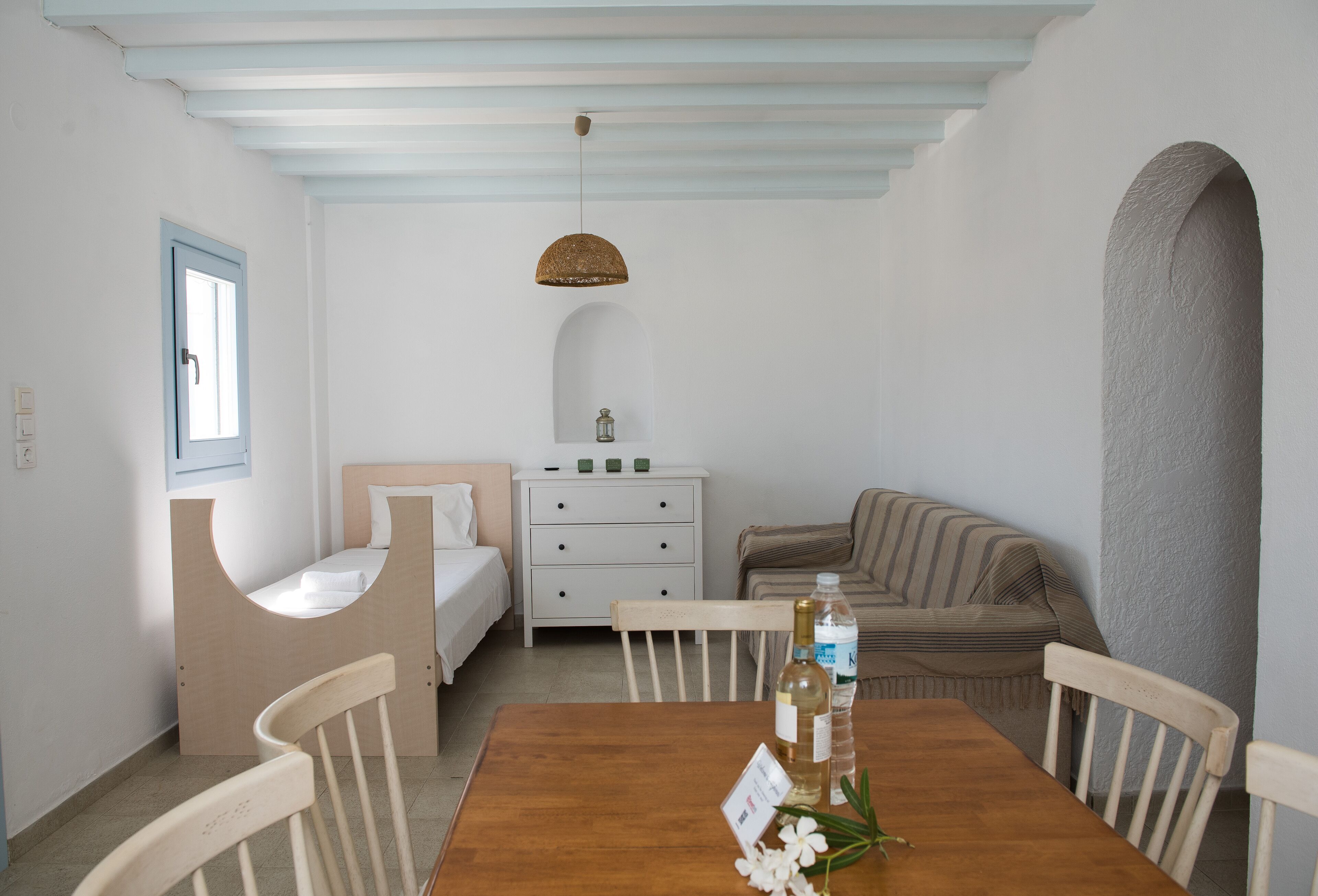 A cozy room with a wooden dining table, chairs, a bed, a nightstand, and a window with a blue frame.