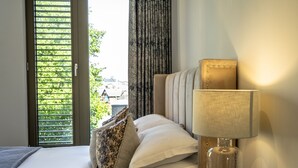 A hotel room with a bed, a lamp, and a view of greenery outside. - A premium stay that sleeps 5 guests  in 2 bedrooms (York)