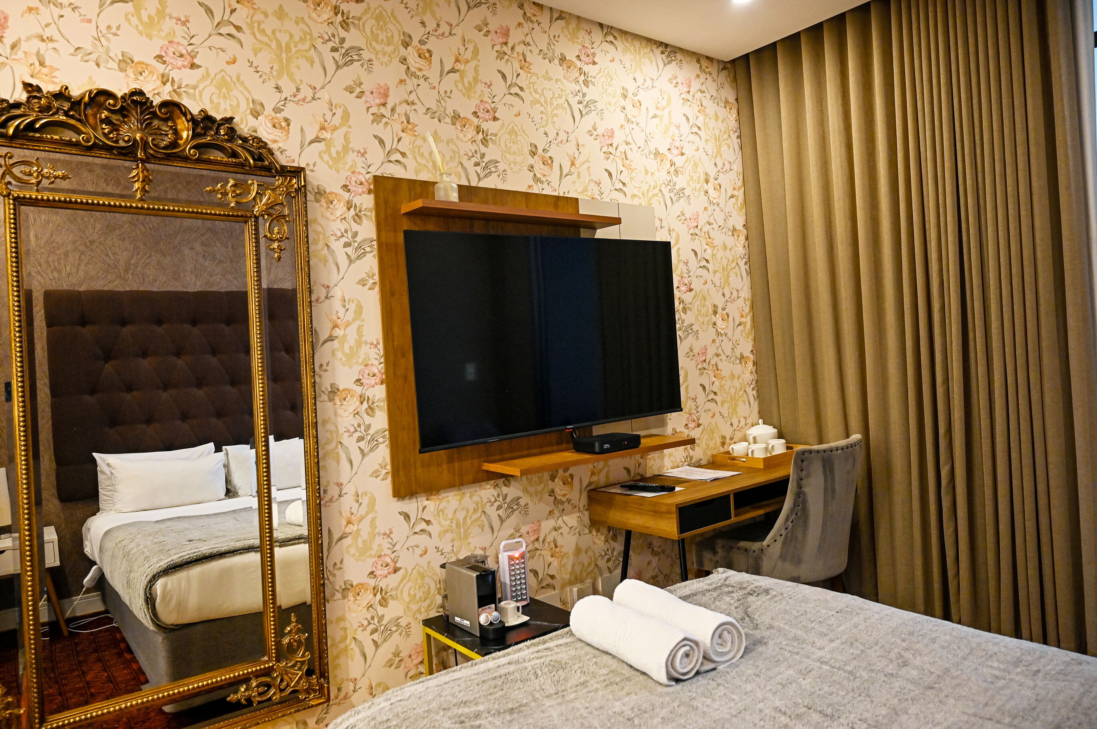 A hotel room with a large mirror, a bed with white linens, a television mounted on the wall, a desk with a chair, and a window with curtains.