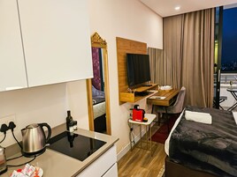 Habitación de hotel moderna con kitchenette, una cama, un escritorio con televisor y vista a la ciudad de noche.