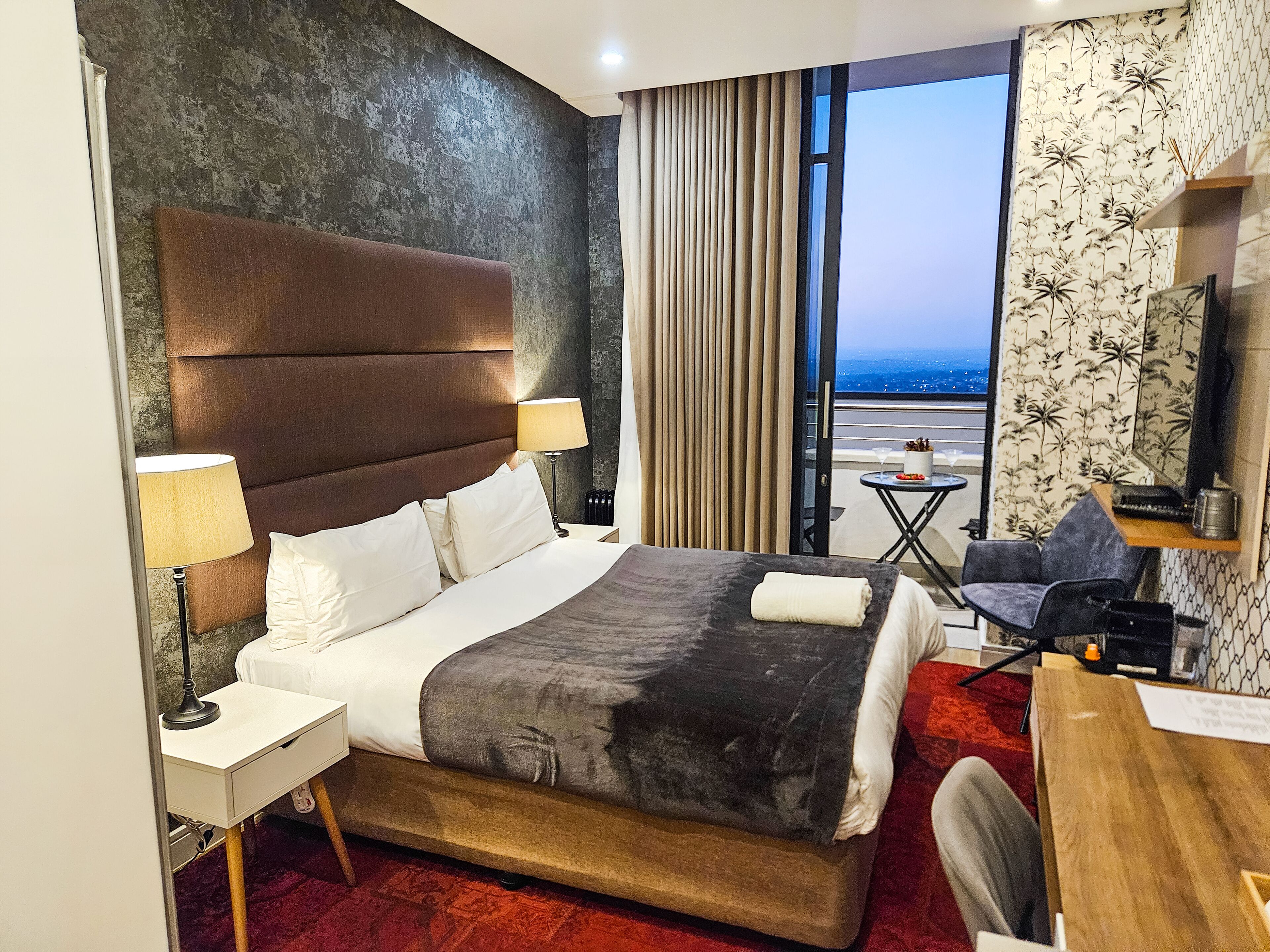 A hotel room with a large bed, a desk, a chair, a nightstand, a lamp, a view of the city, and a window with curtains.