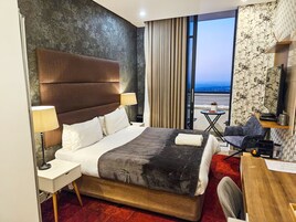 A hotel room with a large bed, a desk, a chair, a nightstand, a lamp, a view of the city, and a window with curtains.
