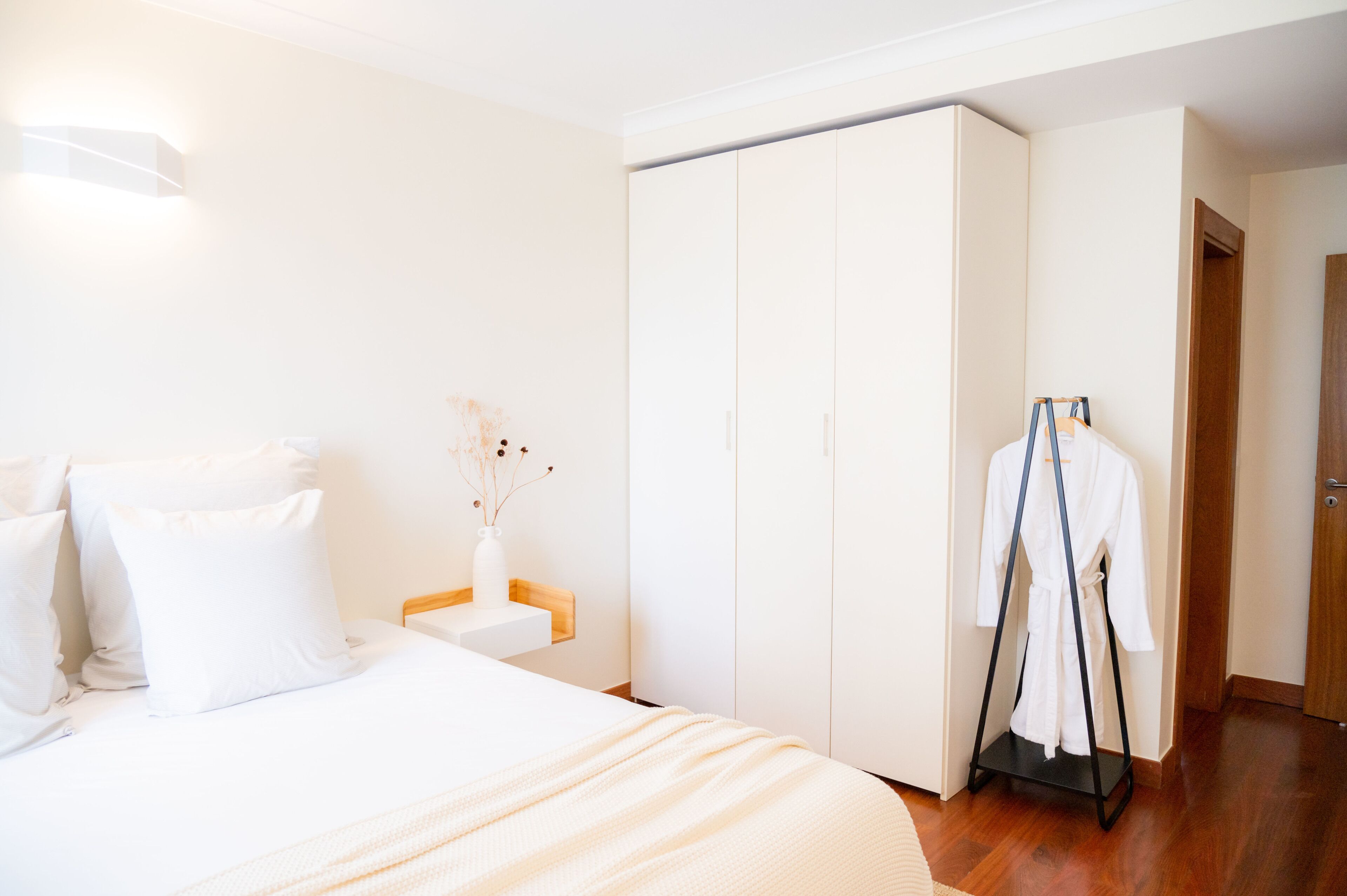 A hotel room with a bed, a wardrobe, a bedside table with a vase, and a robe hanger.