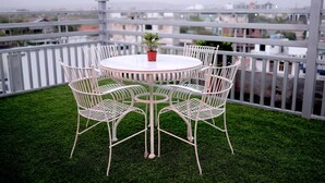 A round table with six white chairs and a potted plant in the center, surrounded by a white railing and green artificial grass. - Hotel SD Prime (Jaipur)