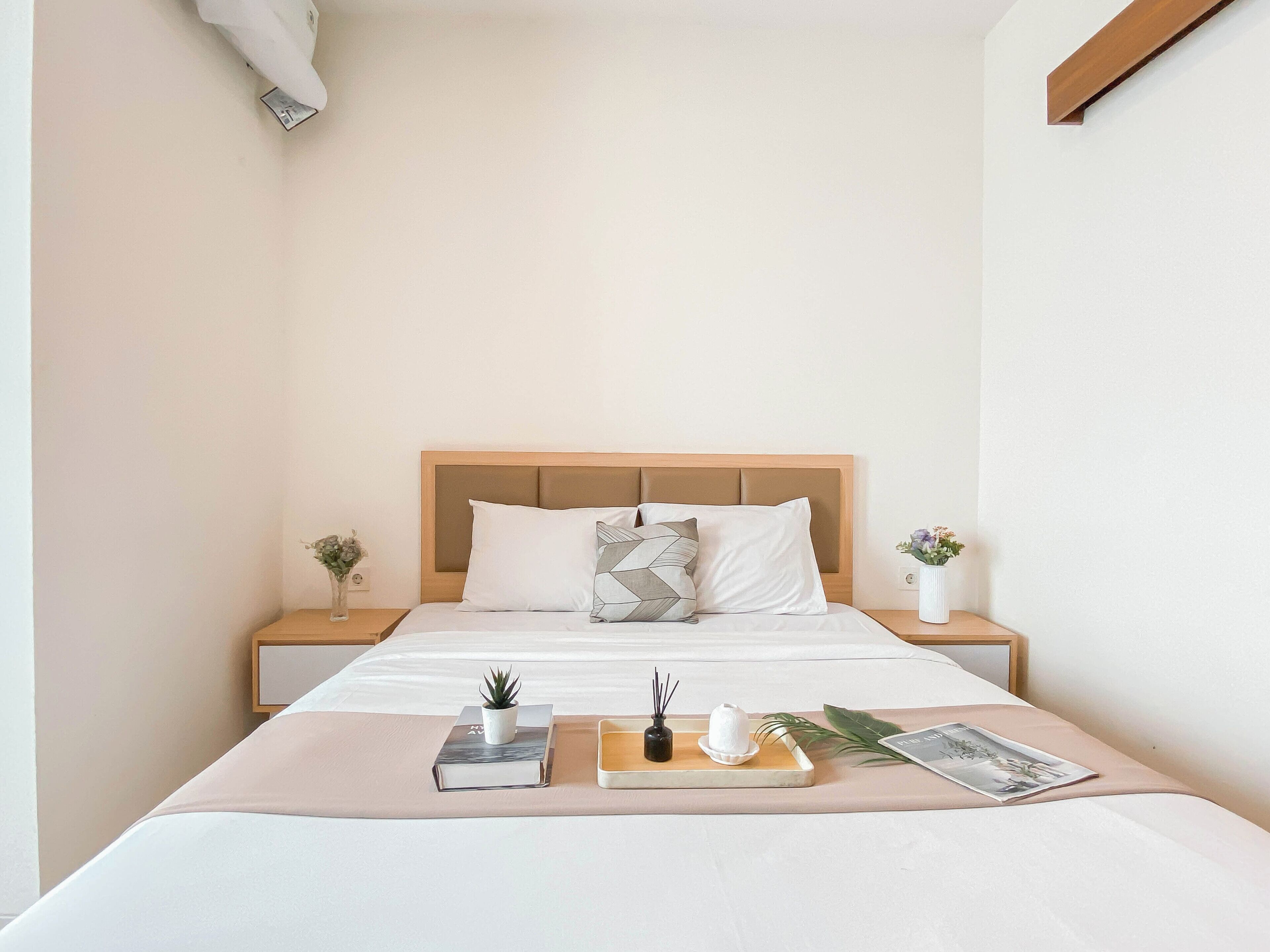 A neatly made bed with a wooden headboard, two bedside tables with small plants, and a bedside table with a book and a small potted plant.
