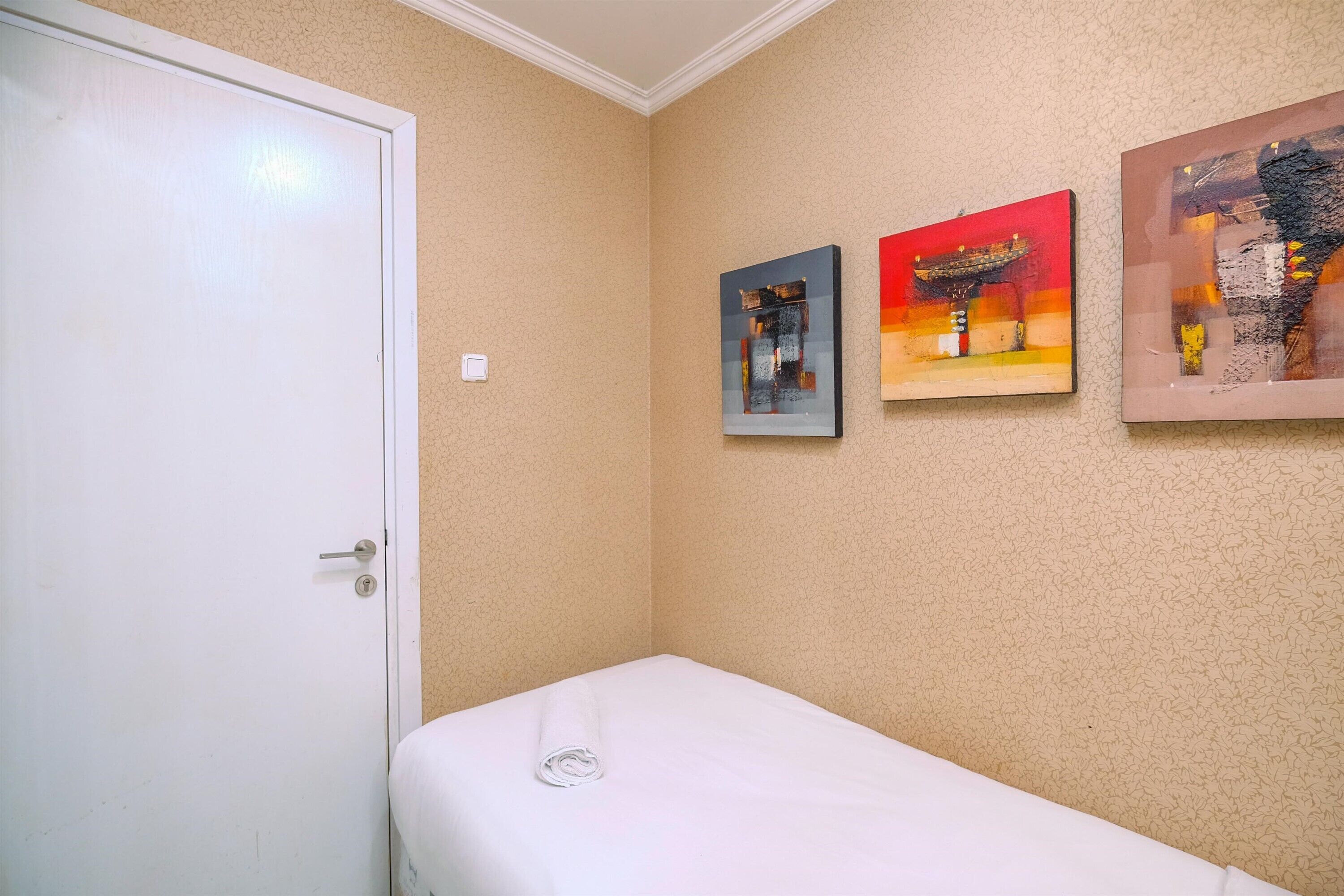 A small, neatly made bed with a white sheet and a rolled towel, a white door, and two framed artworks on the wall.