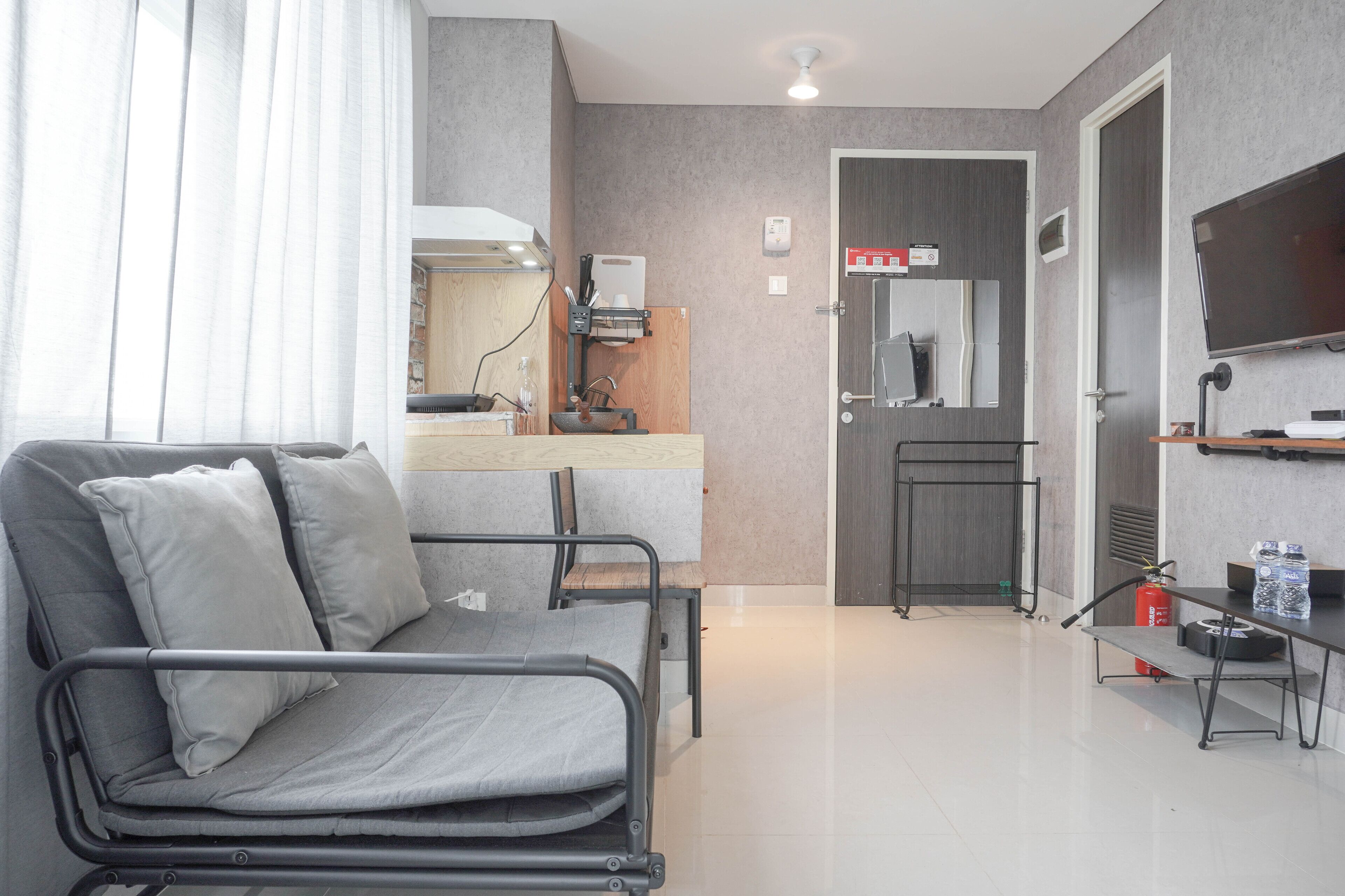 A modern hotel room with a grey sofa, a small kitchenette, a flat-screen TV, and a door leading to a bathroom.