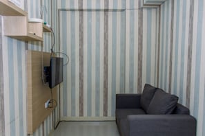 Um quarto pequeno com papel de parede listrado, uma prateleira de madeira com uma TV e um sofá cinza.