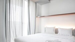Een hotelkamer met een groot bed, witte beddengoed, een net opgemaakt bed met een gevouwen handdoek en dunne gordijnen.