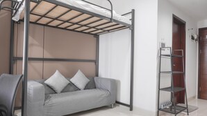 Een stapelbed met een matras, een grijze bank met kussens, een metalen rek en een deur op de achtergrond.
