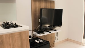 A modern kitchen with a built-in gas stove, a flat-screen TV mounted on a wooden cabinet, and a small black suitcase on the floor. - Great Location Studio Room At Springlake Summarecon Bekasi Apartment (Bekasi)