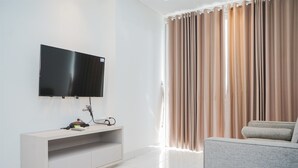 A modern living room with a flat-screen TV mounted on a light-colored console, beige curtains, and a grey sofa. - Comfort And Simple 1Br At Casa De Parco Apartment (South Tangerang)
