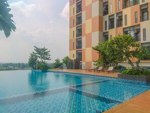 Ein Pool mit gefliesten Rändern, umgeben von Liegestühlen und Grünflächen, neben einem mehrstöckigen Gebäude mit Balkonen.