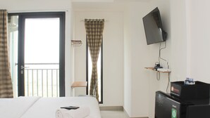 A hotel room with a bed, a TV mounted on the wall, a small desk with a computer, and a window with curtains. - Fancy And Nice Studio Apartment At Sayana Bekasi (Bekasi)