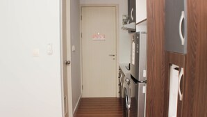 Ein schmaler Flur mit Holzboden, der zu einer Tür führt. Auf der rechten Seite befinden sich Geräte wie eine Waschmaschine und ein Kühlschrank.