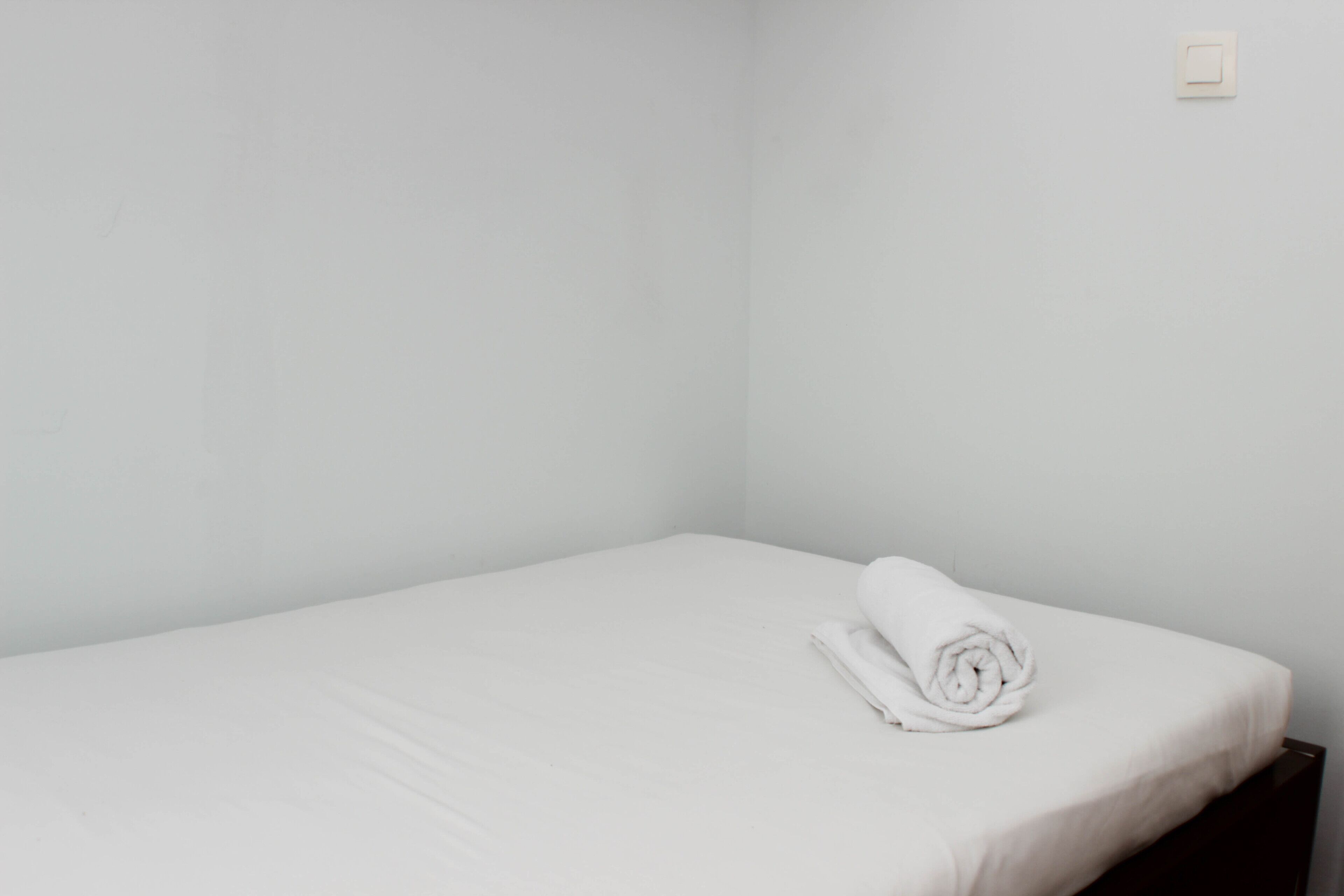 A small, single-bed room with a neatly made bed and a wall-mounted light switch.