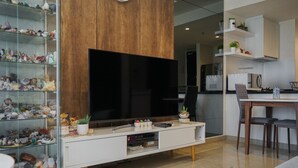 A modern living room with a flat-screen TV, a glass display case with collectibles, a dining area with a wooden table and chairs, and a kitchen in the background. - Minimalist And Cozy 1Br At Branz Bsd City Apartment (Pagedangan)