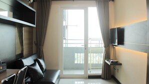 A modern hotel room with a sofa, a flat-screen TV mounted on the wall, a sliding glass door to the balcony, and a small table with a chair. - Comfort 1Br At Sky Terrace Apartment (Jakarta)