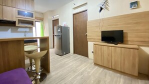 A compact kitchen with wooden cabinets, a built-in refrigerator, a small bar stool, and a television on a wooden stand. - Cozy 3Br Furnished Apartment At Gateway Ahmad Yani Cicadas (Bandung)