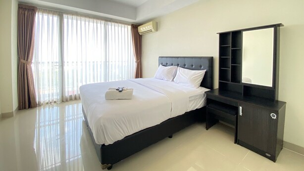A hotel room with a large bed, a bedside table, a mirror, and a window with curtains. - Cozy Studio Apartment At Beverly Dago (Bandung)