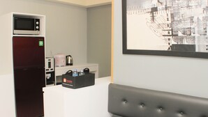 Een compact keukenhoekje met een magnetron, koelkast en koffiezetapparaat. Er is een zithoek met een beklede bank en een kunstwerk in een lijst aan de muur.