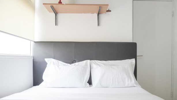 A bed with white bedding and pillows, a grey headboard, and a wooden shelf above. - Best Deal And Comfy 2Br At Green Pramuka City Apartment (Jakarta)