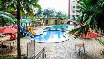 Uma área de recreação infantil com escorregadores e piscina, rodeada por palmeiras e mesas ao ar livre.