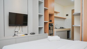 Une chambre d’hôtel moderne dotée d’un téléviseur à écran plat fixé au mur, d’une cuisine intégrée avec un four à micro-ondes et une machine à café, et d’un lit soigneusement fait avec une serviette blanche roulée.