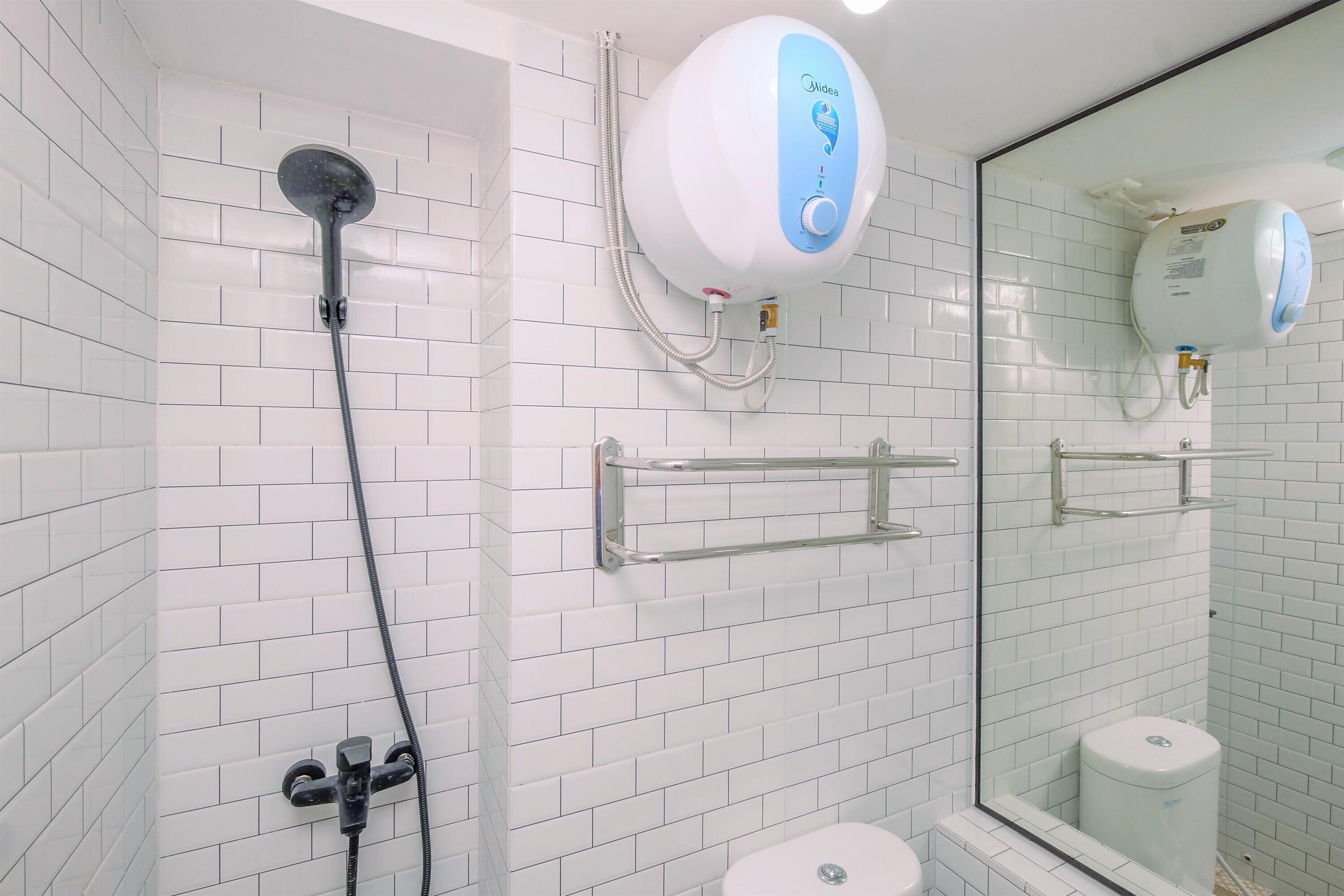 A modern bathroom with white tiled walls, a glass shower enclosure, a handheld showerhead, a toilet, and a water heater mounted on the wall.